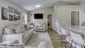 A modern living room with a grey sofa, a white armchair, and a coffee table. There is a flat-screen TV mounted on the wall, a small side table with a vase, and a large window. - Last Mango in Palm Coast (Palm Coast)