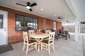 Eine überdachte Terrasse mit einer Ziegelwand, einem runden Holztisch mit Stühlen, Deckenventilatoren und einer Glastür.