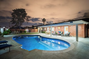 Ein Pool mit gewölbter Kante, eine überdachte Terrasse mit Gartenmöbeln und ein Backsteinhaus mit Tür und Fenstern.