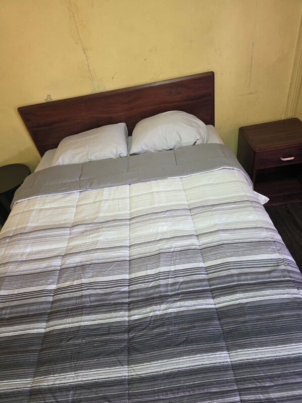 Una cama con colcha a rayas, cabecera de madera, dos almohadas y una mesita de noche con cajón.