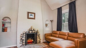 A living room with a stone fireplace, a leather sofa, a bookshelf, and a window with curtains. - Whimsical Historic Getaway Near Golf, Skiing and Wisconsin Southern Lakes (Genoa City)