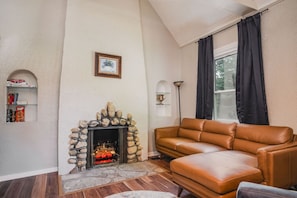 A living room with a stone fireplace, a leather sofa, a bookshelf, and a window with curtains.