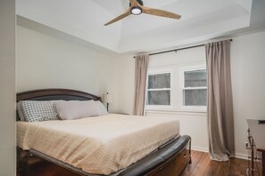 A bedroom with a bed, a ceiling fan, a window with curtains, and a desk with a lamp.