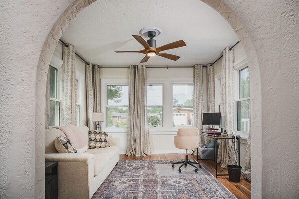 A room with a ceiling fan, a sofa, a desk with a computer, and a window with curtains.