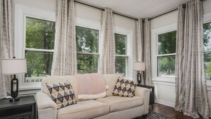 A cozy living room with a beige sofa, patterned cushions, and a large window with curtains. - Whimsical Historic Getaway Near Golf, Skiing and Wisconsin Southern Lakes (Genoa City)