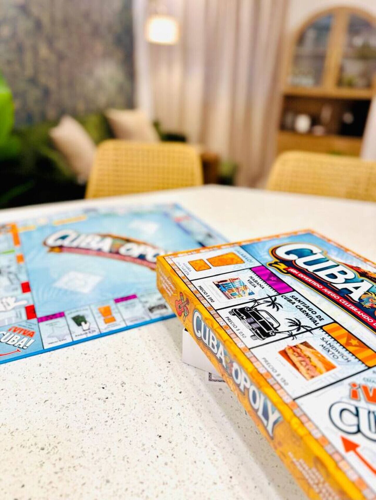 Dos juegos de mesa, Cuba y Cuba Opoly, sobre una mesa en una habitación con sillas de mimbre y una estantería.