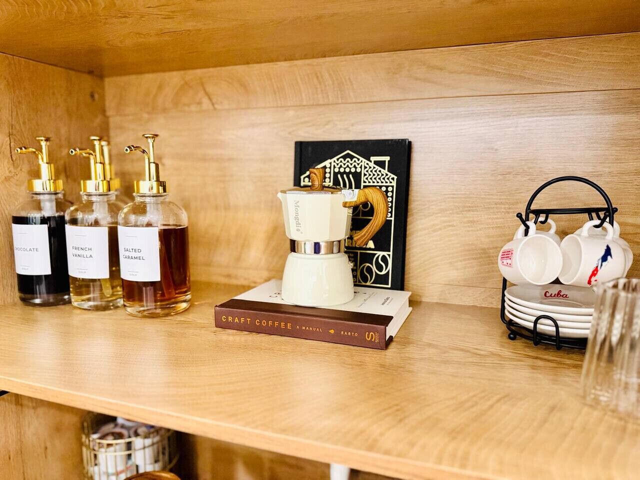 A wooden shelf with coffee-making equipment, bottles of flavored syrups, a coffee manual, and a mug rack.
