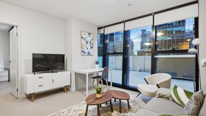 Un salon moderne doté d’un téléviseur à écran plat, d’un bureau blanc, d’un fauteuil confortable, d’un canapé et de grandes fenêtres offrant une vue sur la ville.