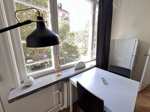Ett litet kök med en vit bänkskiva, en svart lampa, ett vitt element och ett fönster med utsikt över byggnader och träd.