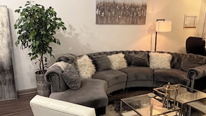 Een moderne woonkamer met een hoekbank, een witte fauteuil, een glazen salontafel en een potplant.