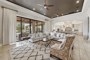 Ein geräumiges Wohnzimmer mit einer großen Eckcouch, einem Couchtisch und einem gemusterten Teppich. Es gibt Deckenventilatoren, eingelassene Beleuchtung und einen Ausblick ins Freie.