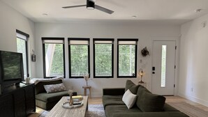 Una sala de estar moderna con un televisor de pantalla plana, dos sofás, una mesa de centro y ventanas amplias.
