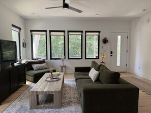 A modern living room with a flat-screen TV, two sofas, a coffee table, and large windows.