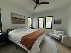 Une chambre à coucher comprenant un lit, un ventilateur de plafond, une table de chevet, une lampe, une chaise et une fenêtre donnant sur l’extérieur.