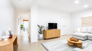 Una sala moderna con un sofá, un televisor sobre un mueble de madera, un comedor con una mesa y un pasillo que conduce a una puerta.