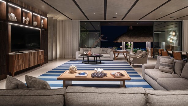 A modern living room with a large flat-screen TV, a wooden entertainment unit, a striped rug, and a dining area with a thatched ceiling. - Casa De Elementos in Punta Mita (Punta de Mita)