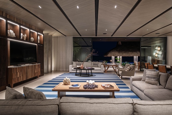 A modern living room with a large flat-screen TV, a wooden entertainment unit, a striped rug, and a dining area with a thatched ceiling. - Casa De Elementos in Punta Mita (Punta de Mita)