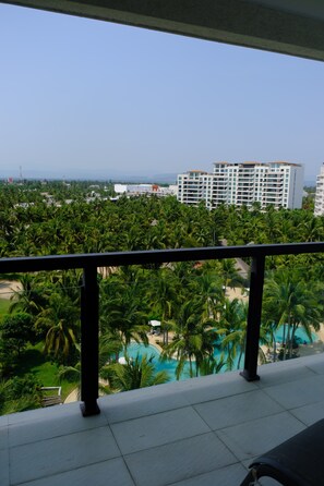 A balcony with a view of a swimming pool surrounded by palm trees and a distant building. - Acapulco Diamante Apartment (pet friendly) (Acapulco de Juárez)