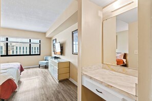 A bedroom with a bed, a dresser, a chair, a desk, a TV, and a large mirror. - Oceanview Coastal Retreat | Heated Pool, Hot Tub, & Steps to the Beach! 🍹 (Cocoa Beach)