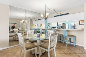 A dining area with a glass table surrounded by white chairs, a kitchen with white cabinets, and a blue countertop. - Oceanview Coastal Retreat | Heated Pool, Hot Tub, & Steps to the Beach! 🍹 (Cocoa Beach)