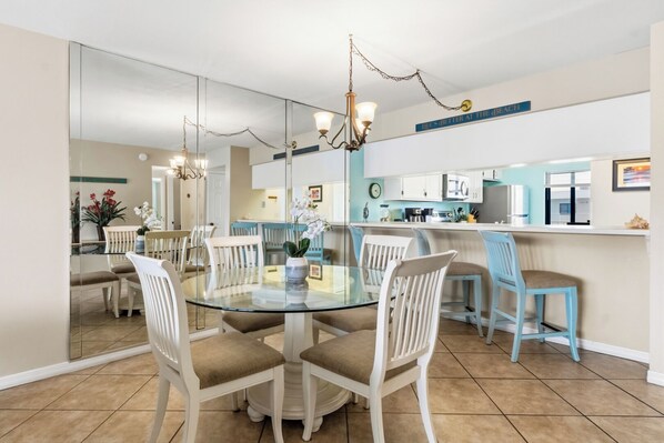 A dining area with a glass table surrounded by white chairs, a kitchen with white cabinets, and a blue countertop. - Oceanview Coastal Retreat | Heated Pool, Hot Tub, & Steps to the Beach! 🍹 (Cocoa Beach)