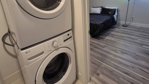 Un área de lavandería con lavadoras y secadoras apiladas, una cama con ropa de cama estampada, una lámpara de pie y un suelo de madera.