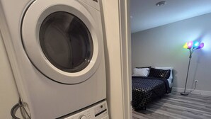 Un área de lavandería con lavadoras y secadoras apiladas, una cama con ropa de cama estampada y una lámpara de pie.