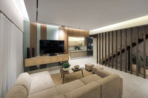 Ein modernes Wohnzimmer mit Sofa, Couchtisch und Fernseher, das in eine Küche und eine Treppe übergeht.