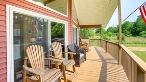 A covered deck with wooden chairs and a table, a sliding glass door, and a view of greenery. - Island Gem, Private Yard, Fire Pit, Patio Dining, Pellet Smoker and Gas Grills (Kelleys Island)