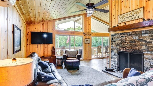 A cabin interior with a fireplace, wooden walls, a ceiling fan, a television, and a seating area. - Island Gem, Private Yard, Fire Pit, Patio Dining, Pellet Smoker and Gas Grills (Kelleys Island)