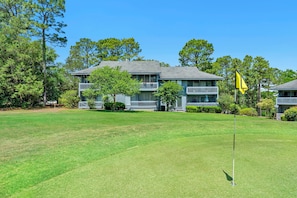 Ein Golfplatz mit Fahnenstange, ein Gebäude mit Balkonen und umliegende Bäume.
