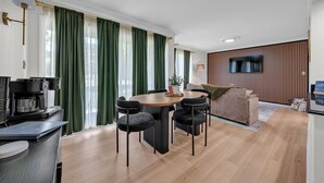 A modern living room with a round table, four chairs, a sofa, a TV mounted on the wall, and a coffee machine.