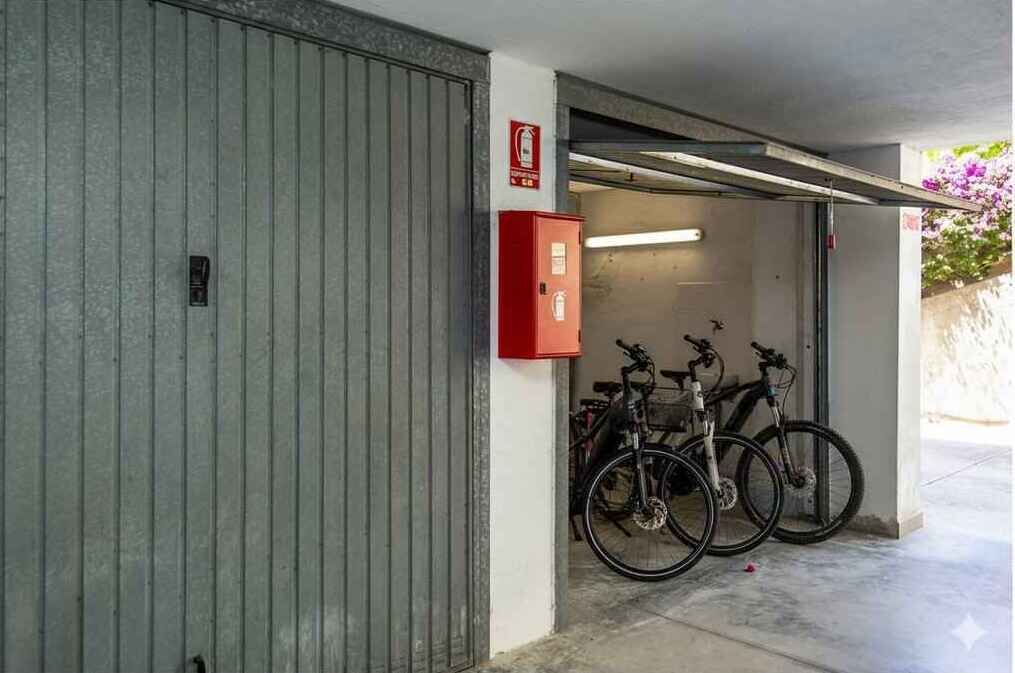 A garage with a closed metal door, a red fire alarm box, and two bicycles parked inside. - Holiday apartment with air conditioning and balcony (Piani Paorelli)