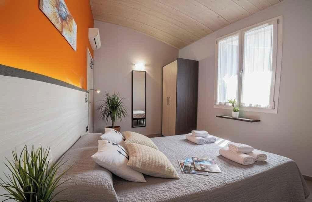Un dormitorio con cama, almohadas, una mesita de noche, un espejo, una ventana y una planta.