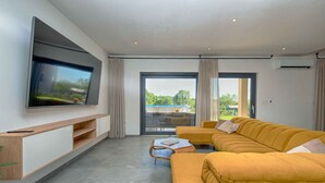 Ein modernes Wohnzimmer mit einem großen Flachbildfernseher, einem gelben Ecksofa, einem Couchtisch und einer Glasschiebetür, die zu einem Außenbereich führt.