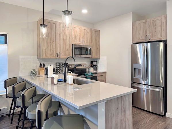 A modern kitchen with wooden cabinets, stainless steel appliances, a central island with bar stools, and pendant lighting. - Basecamp - Ski·Golf·Fish·Lake - Fire Table + Views (Granby)