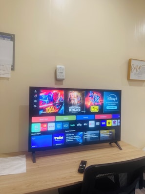 A flat-screen TV mounted on a wall with a remote control on a wooden desk.