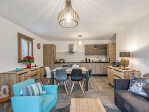Eine moderne Küche mit Holzschränken, einem Esstisch mit Stühlen und einer zentralen Kochinsel. Im Wohnbereich befindet sich eine Couch und ein Couchtisch.