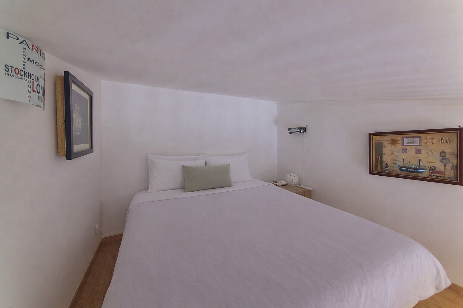 Une chambre avec un lit simple, une table de chevet, un tableau encadré au mur et un panneau indiquant des noms de villes.