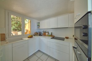 A modern kitchen with white cabinets, a built-in oven, and a window with a view of greenery. - Villa Stretta d’A Pianedda – 180° Ocean View - Welkeys (Villanova)