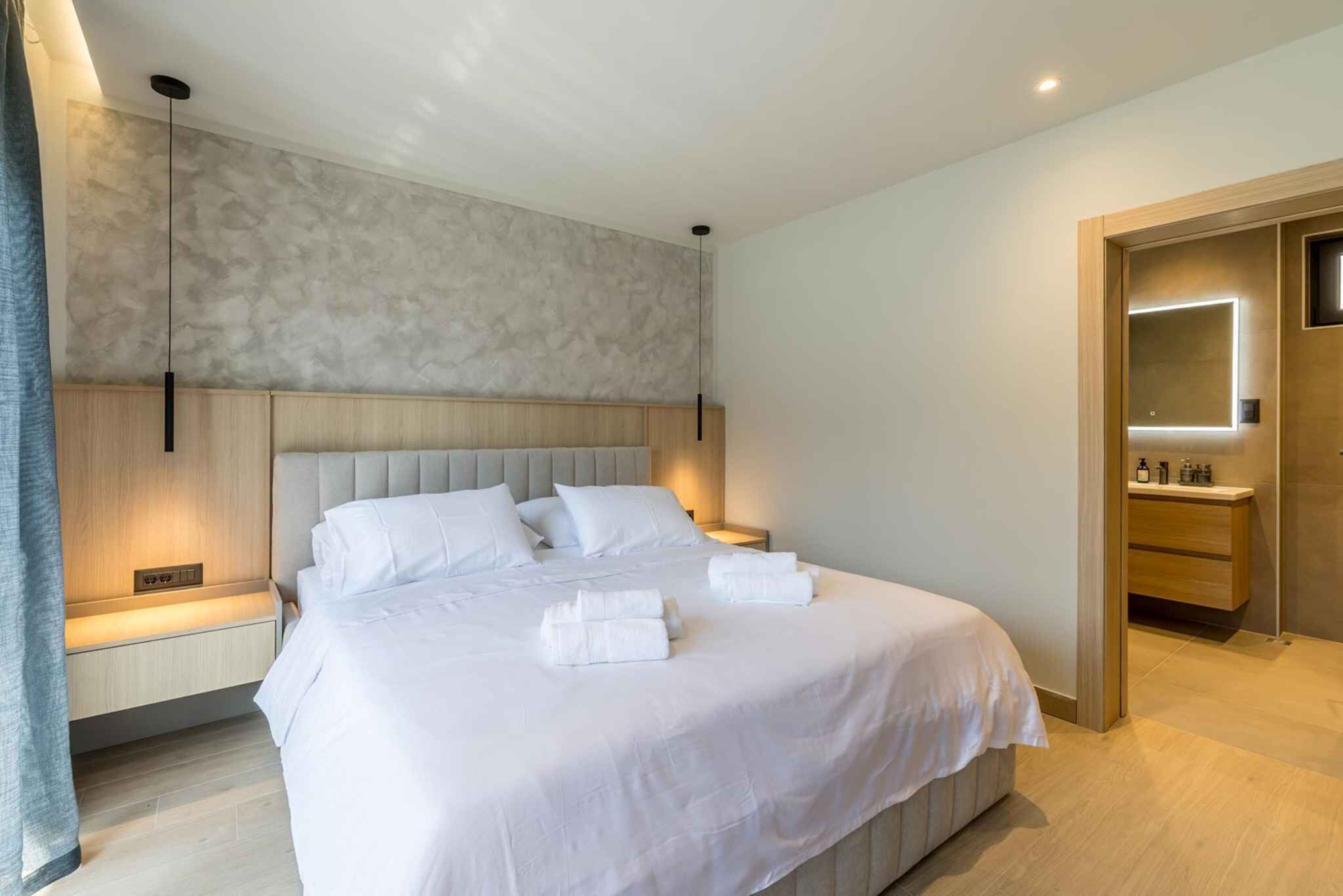 A modern hotel room with a large bed, bedside tables, a built-in bench, a wooden door, and a bathroom with a mirror and sink.