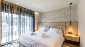 Moderni hotellihuone, jossa on suuri sänky, yöpöytä, seinään kiinnitetty lamppu ja näkymä ohuiden verhojen läpi.
