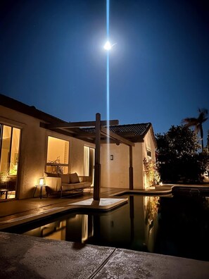Ein Haus bei Nacht mit Pool, einer Terrasse mit Sofa und einer hellen Lichtquelle darüber.