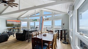 A spacious living area with a high ceiling, wooden dining table, comfortable seating, and large windows offering views of the outdoors. - Salty Oyster | Waterfront living at it (Chincoteague Island)