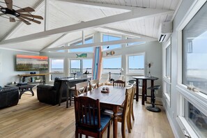 Een ruime woonkamer met een hoge plafond, een houten eettafel, zachte zitmogelijkheden en grote ramen met uitzicht op het landschap.