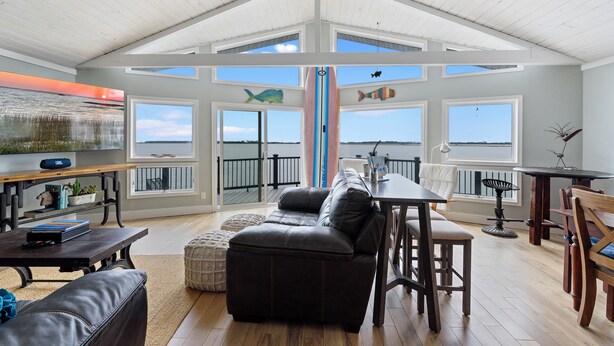 A spacious living area with a high ceiling, large windows, a black leather sofa, a wooden dining table, and a view of the water. - Salty Oyster | Waterfront living at it (Chincoteague Island)