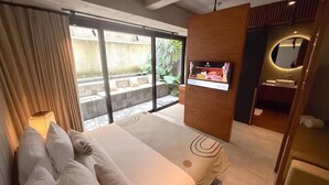 Una camera d'albergo moderna con un letto grande, TV integrata, bagno piccolo e una porta scorrevole in vetro che dà all'esterno.