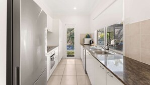 A narrow kitchen with white cabinets, a stainless steel refrigerator, and a window overlooking a garden. - Coastal 4-Bed Retreat with Pool in Toogoom (Toogoom)