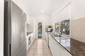 A narrow kitchen with white cabinets, a stainless steel refrigerator, and a window overlooking a garden. - Coastal 4-Bed Retreat with Pool in Toogoom (Toogoom)