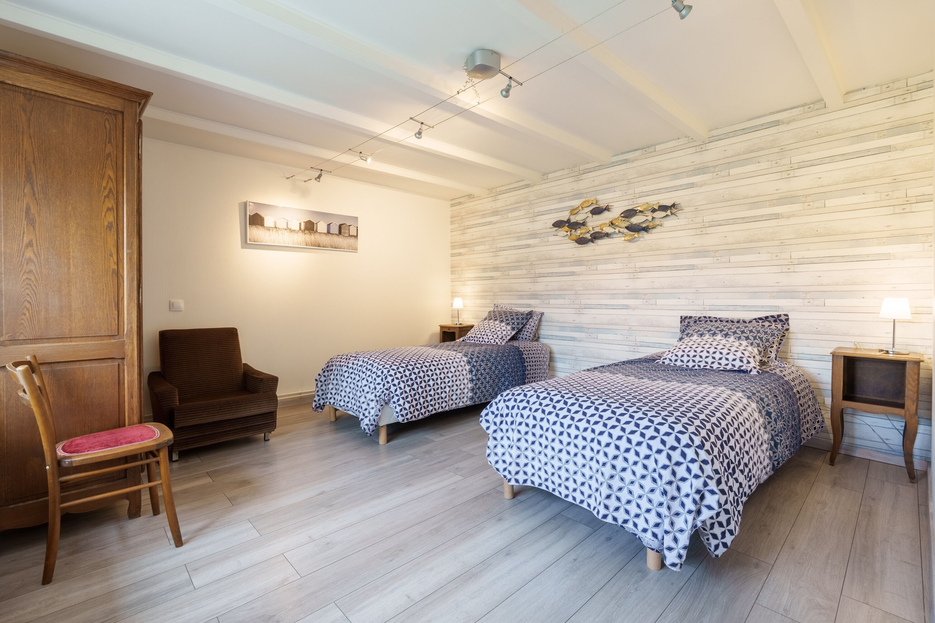 A bedroom with two beds, a wooden wardrobe, a chair, and a wall-mounted light fixture.